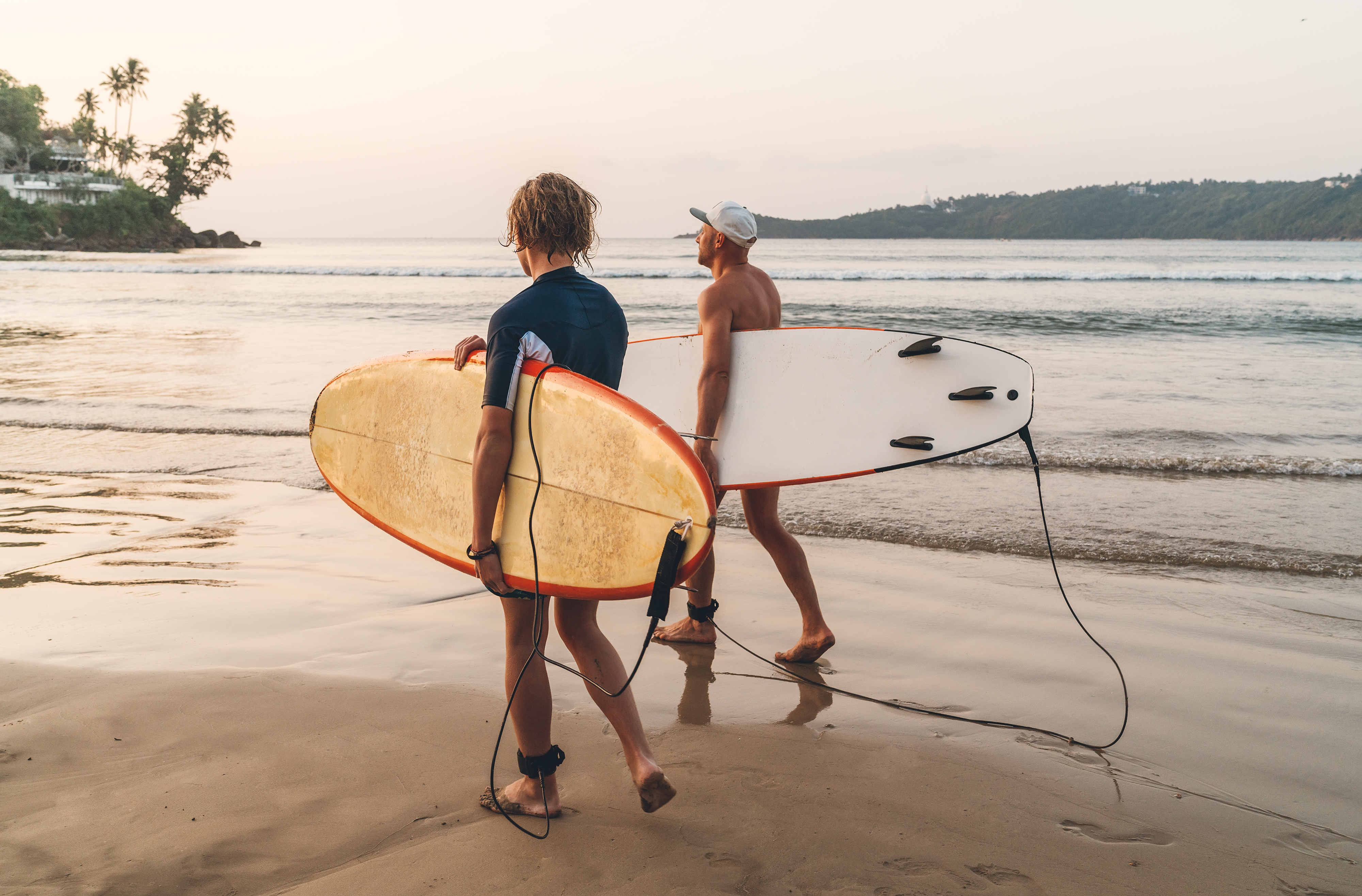 Two individuals stroll along the beach, each carrying a surfboard under their arms, enjoying the coastal scenery.